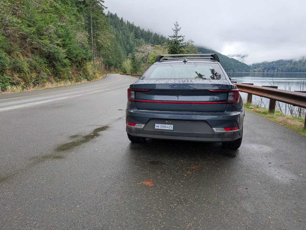 The back of a dark blue car parked next to water with a pine covered hill on the other side.