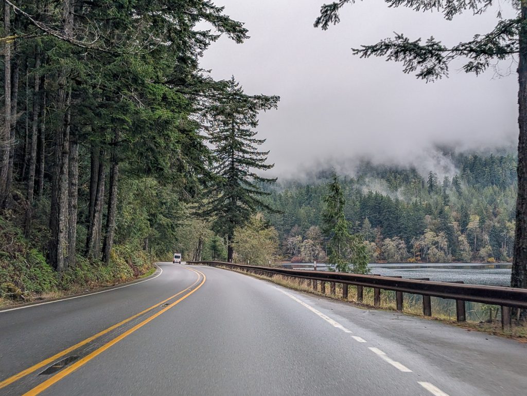 A road with a white van in the distance, pine trees on one side and water on the other, with mist covered hill leading down to the water in the background.