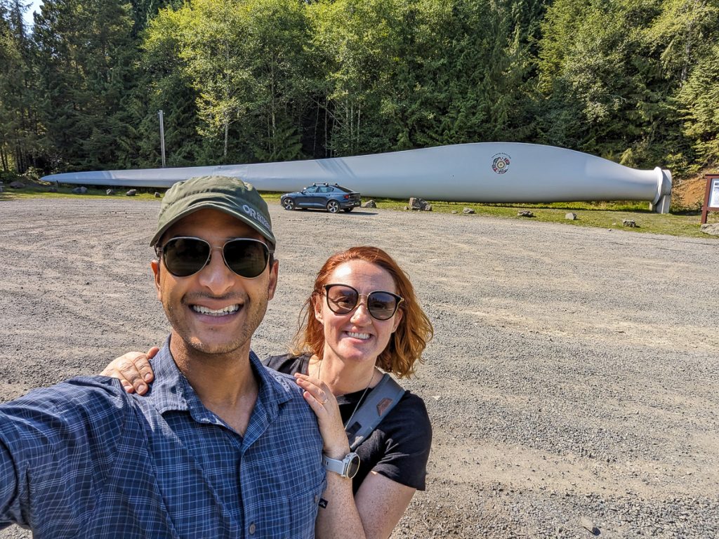 A man and woman smiling for a selfie with a dark blue car, a wind turbine and pine trees in the background.