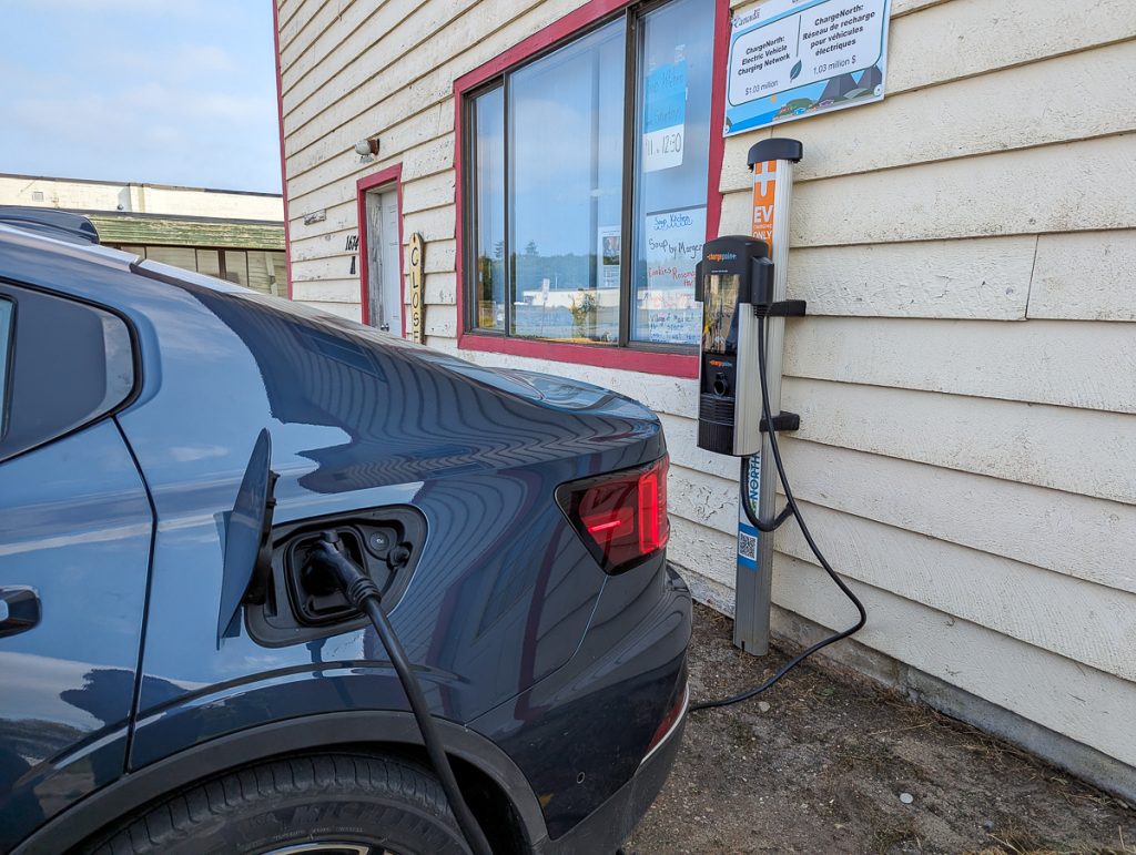The end of a dark blue car plugged into an electric vehicle charger on a weatherboard building.