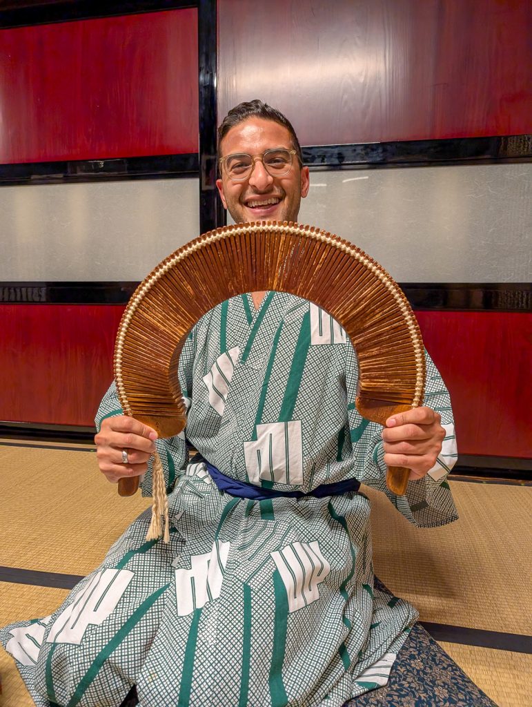 Ganesh kneeling on the floor in a Japanese robe while grinning at the camera and holding a half-circle shaped wooden object.