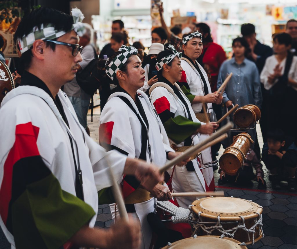 A row of Japanese Taiko drummers in traditional dress on a busy street.