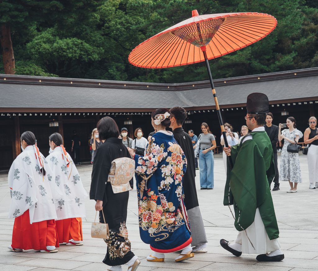 Six Japanese people in traditional wedding dress walking under a parasol with people in the background watching and taking photos.