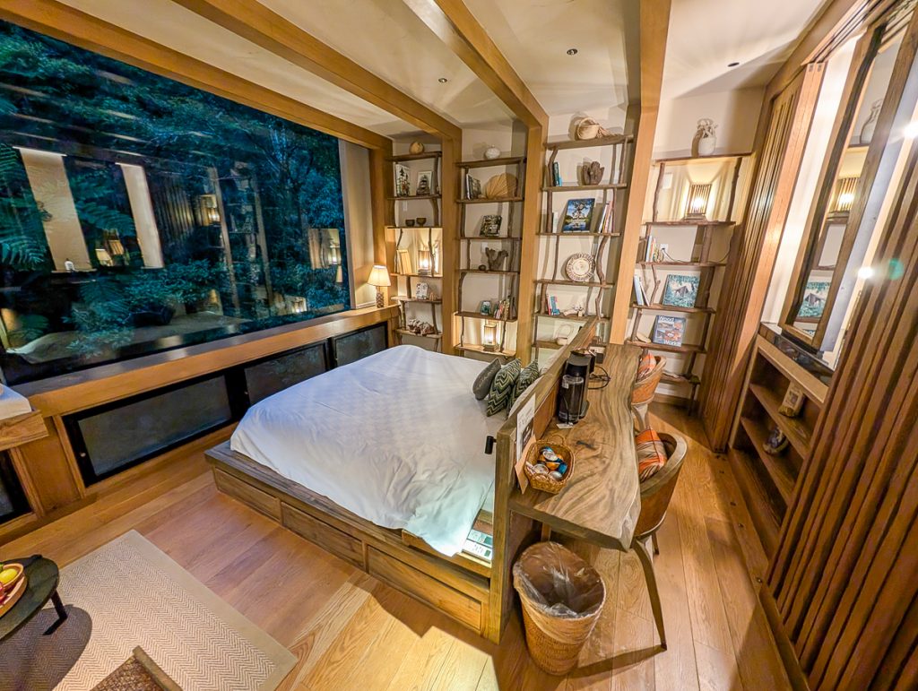 A luxurious hotel room with lots of wood accents and shelves plus a bed in the middle of the room facing a window showing nothing but greenery outside.
