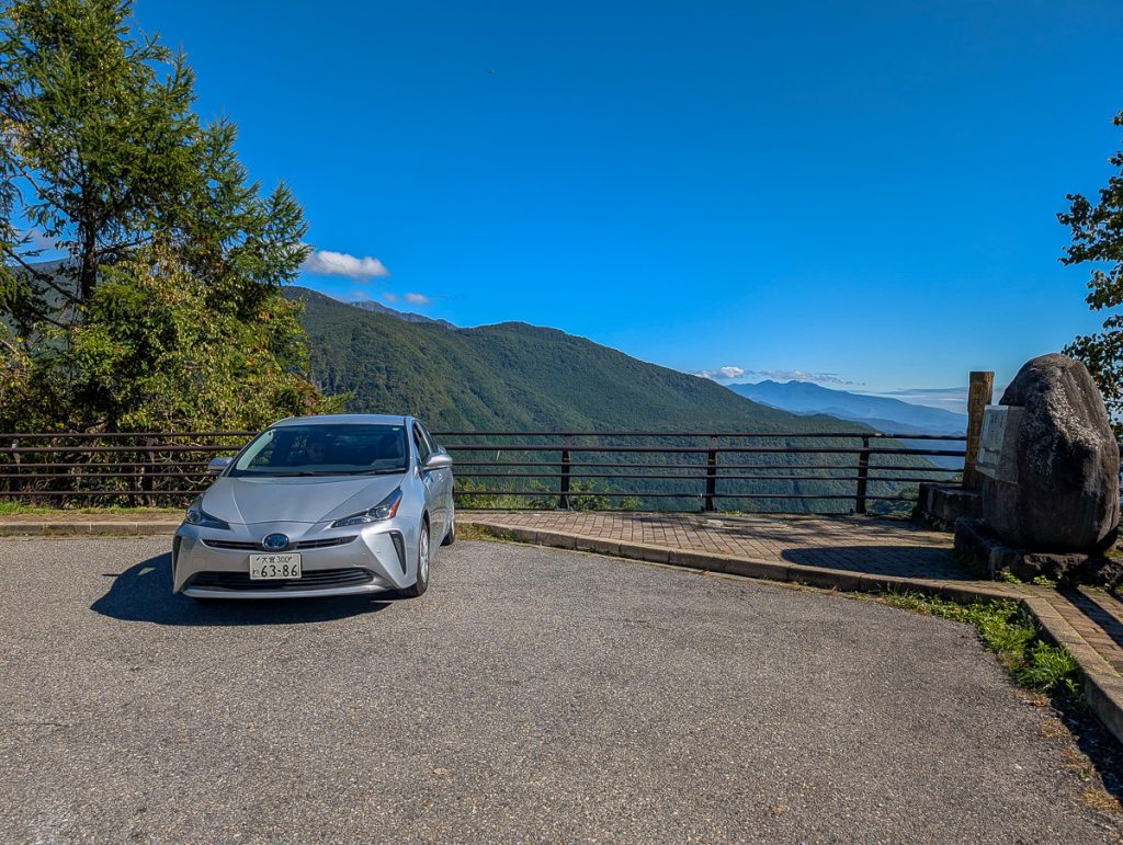 A silver car with a Japanese number plate parked next to a fence looking out over forested mountains and a clear blue sky with just a couple of fluffy white clouds.