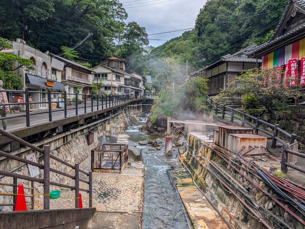 A creek with steam coming off it next to stairs leading up to a street, traditional Japanese houses and greenery in the background.