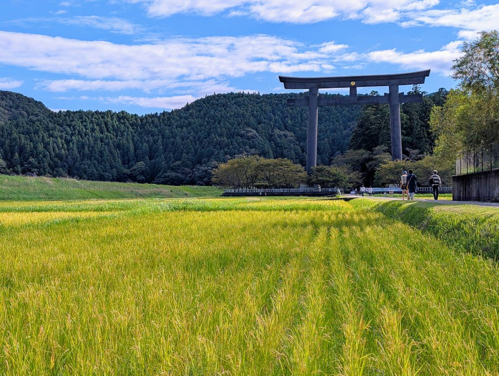 A field of green grass leading to a large Tori Gate with people walking on a path towards it and forested hills behind it.