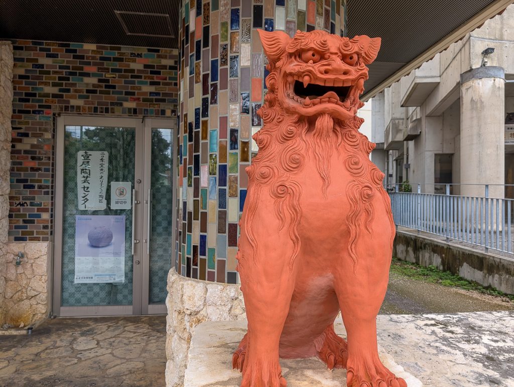 A reddish statue of a lion-dog in Okinawa, sitting on front of a building with different colored mosaic bricks.