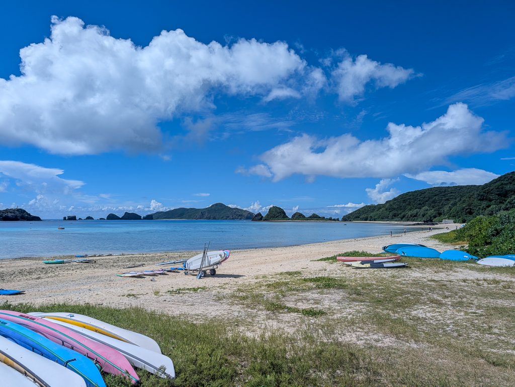 Grass leading to a beach with lots of kayaks and paddleboards lying around. In the background are small green islands, blue sky and fluffy white clouds.