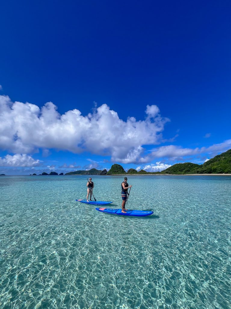 A man and woman on stand up paddleboards in crystal clear water with some green land behind them, fluffy white clouds and blue sky.
