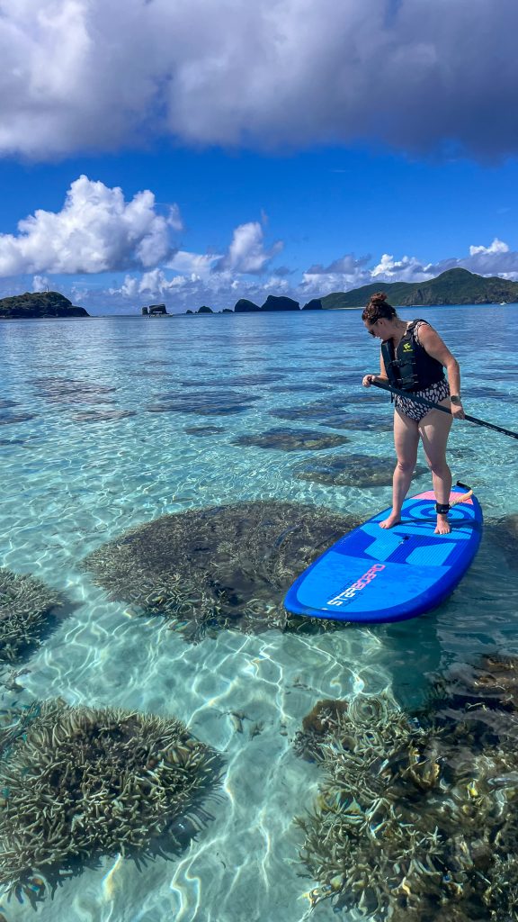 Sooz on a stand up paddleboard looking down through the water at coral with blue sky and fluffy white clouds in the background.