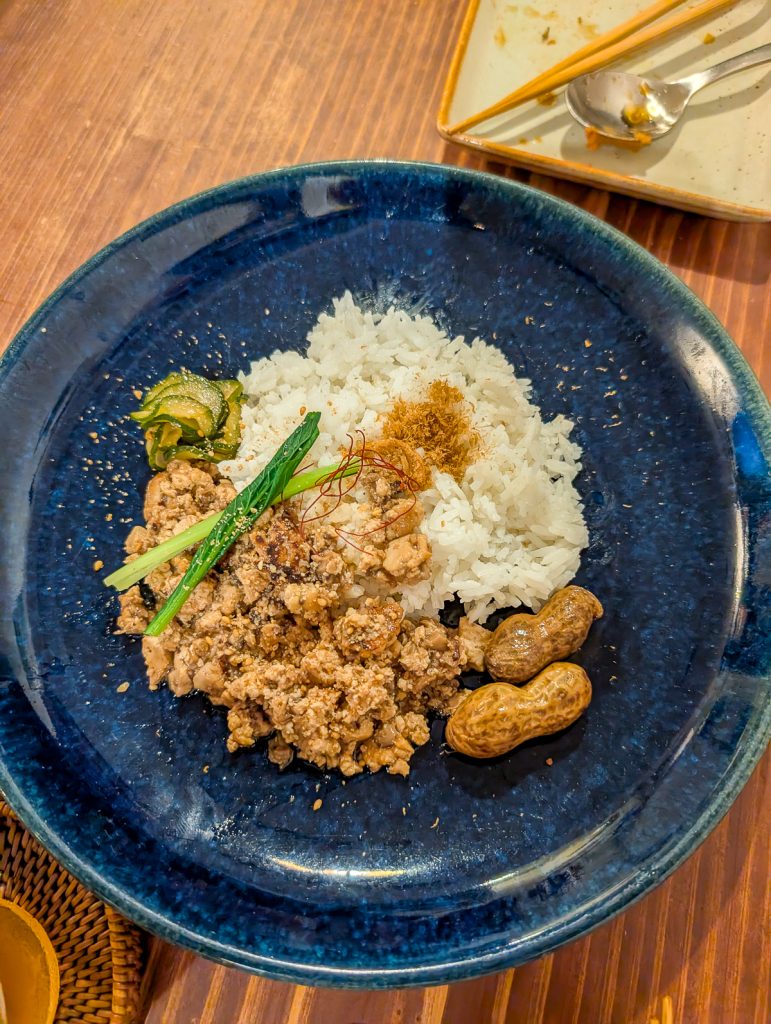 A blue plate holding some white rice, two whole peanuts and some kind of brown food with green garnishes.