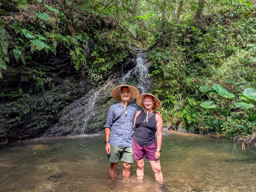 Sooz and Ganesh posing with their arms around each other while wearing straw hats and standing in water with a small waterfall and greenery behind them.