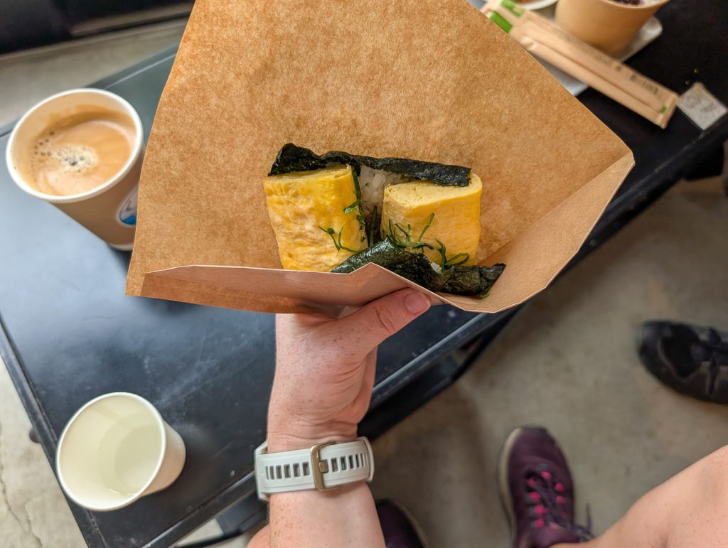 A hand holding a seaweed, rice and tofu snack in brown paper, with a cup of coffee on a black surface below it.