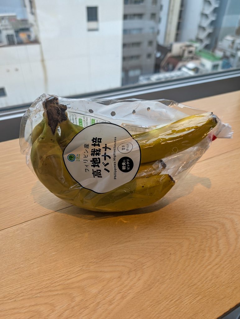 A bunch of bananas in plastic with Japanese text on the plastic, sitting on a wooden surface with a view of buildings out the window behind it.