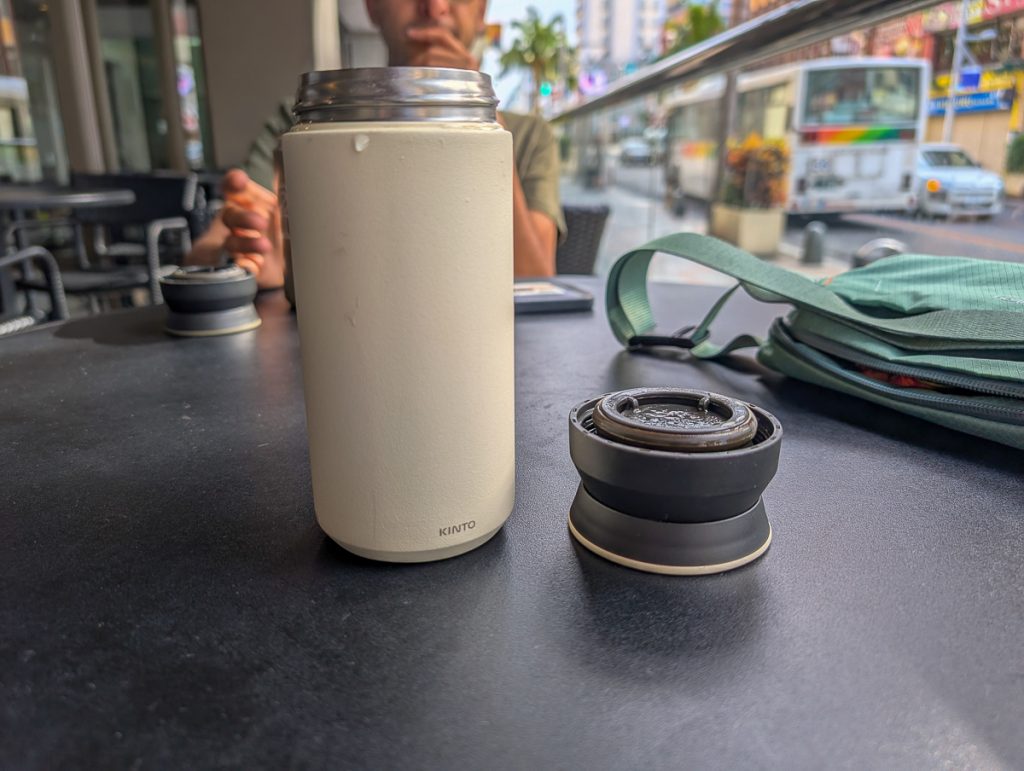 A pale beige reusable drink bottle with Kinto written on it sitting on a table next to the lid and a backpack.