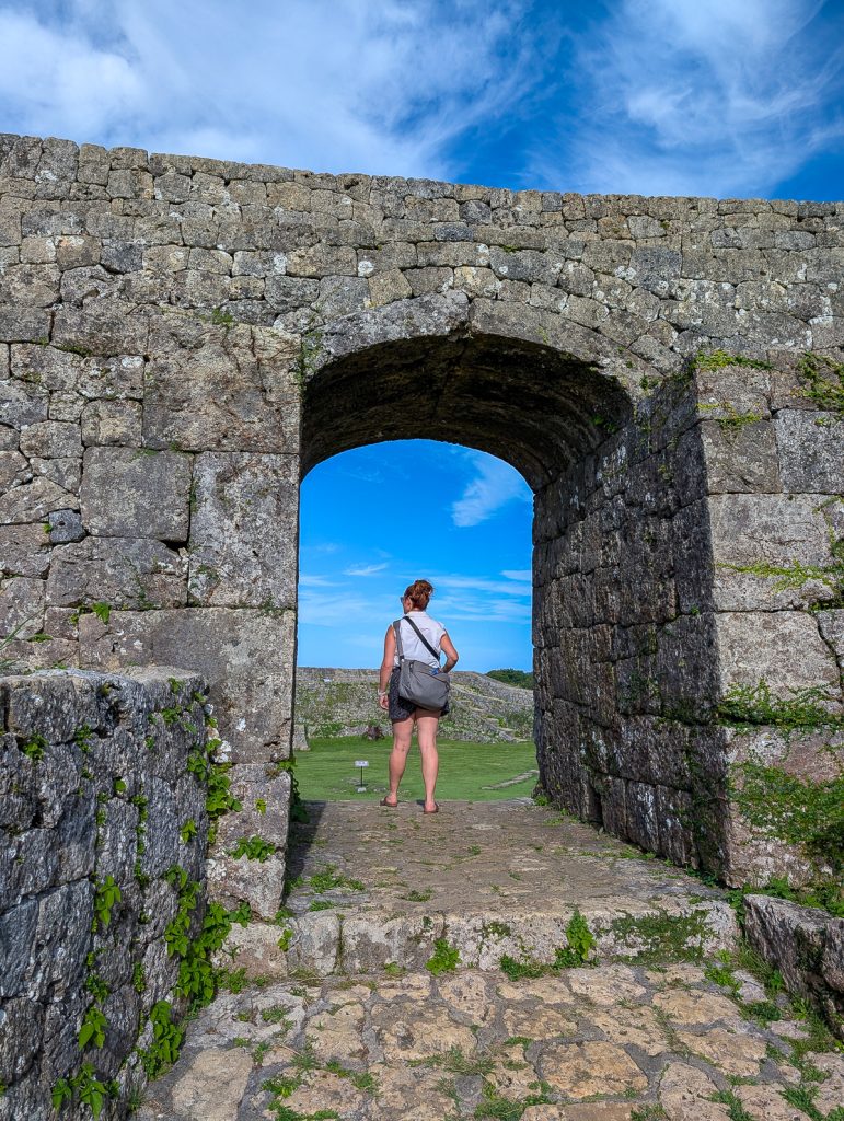 A woman standing with her back to the camera under an archway in a stone wall.