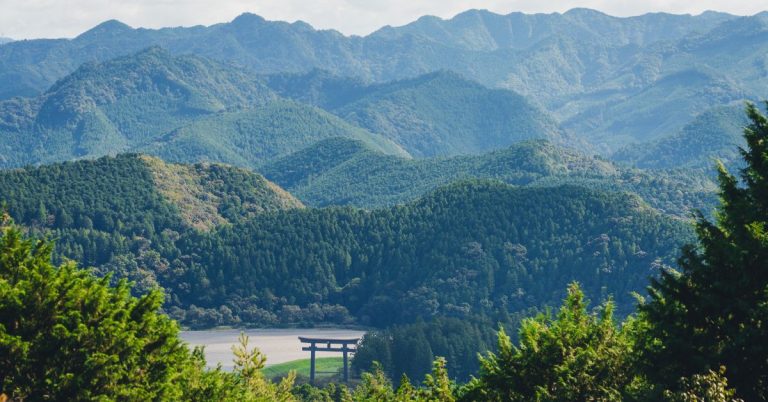 A landscape of forested mountains and a large Japanese wooden gate at the bottom of a valley.
