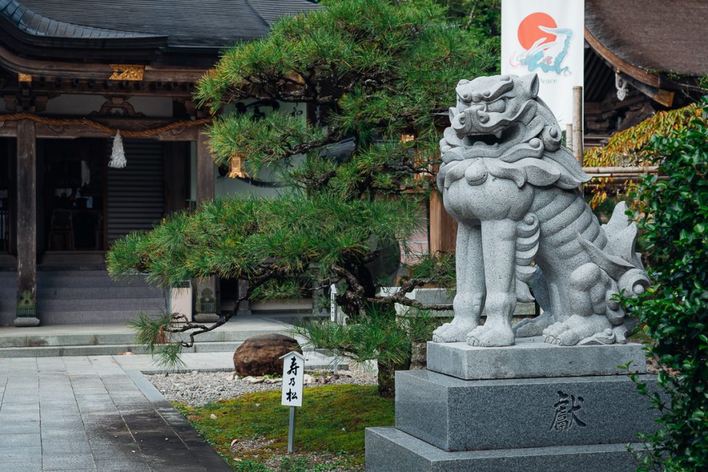 A statue of a lion-dog outside a traditional Japanese building with ornamental trees and signs around.