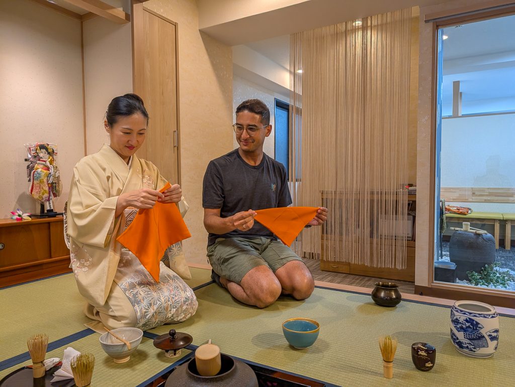 Ganesh and a Japanese woman in a kimono kneeling on a tatami mat and folding orange cloths with tea ceremony utensils on the floor in front of them.