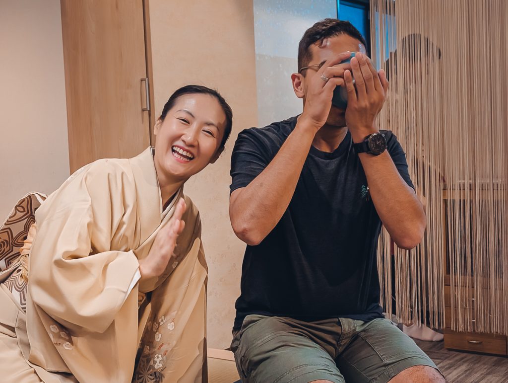 Ganesh drinking tea with his hands and cup covering his face while a Japanese woman in a kimono next to him smiles and waves at the camera.
