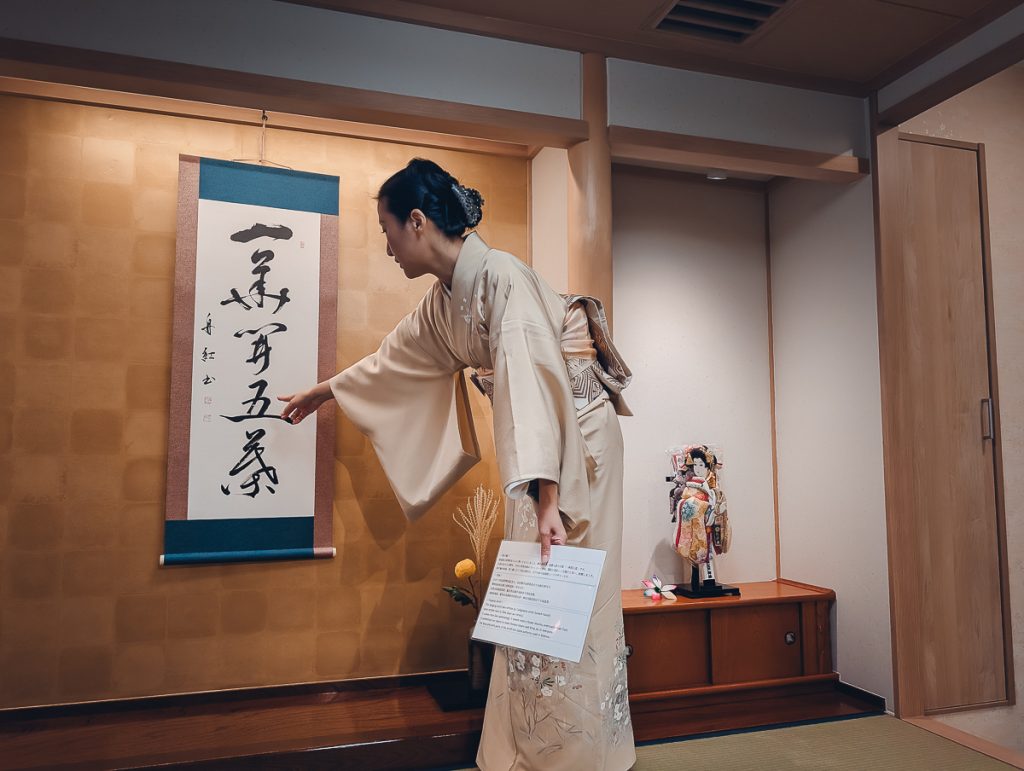 A Japanese woman in a kimono holding a sheet of paper and pointing at a poster on a wall with Japanese writing on it.