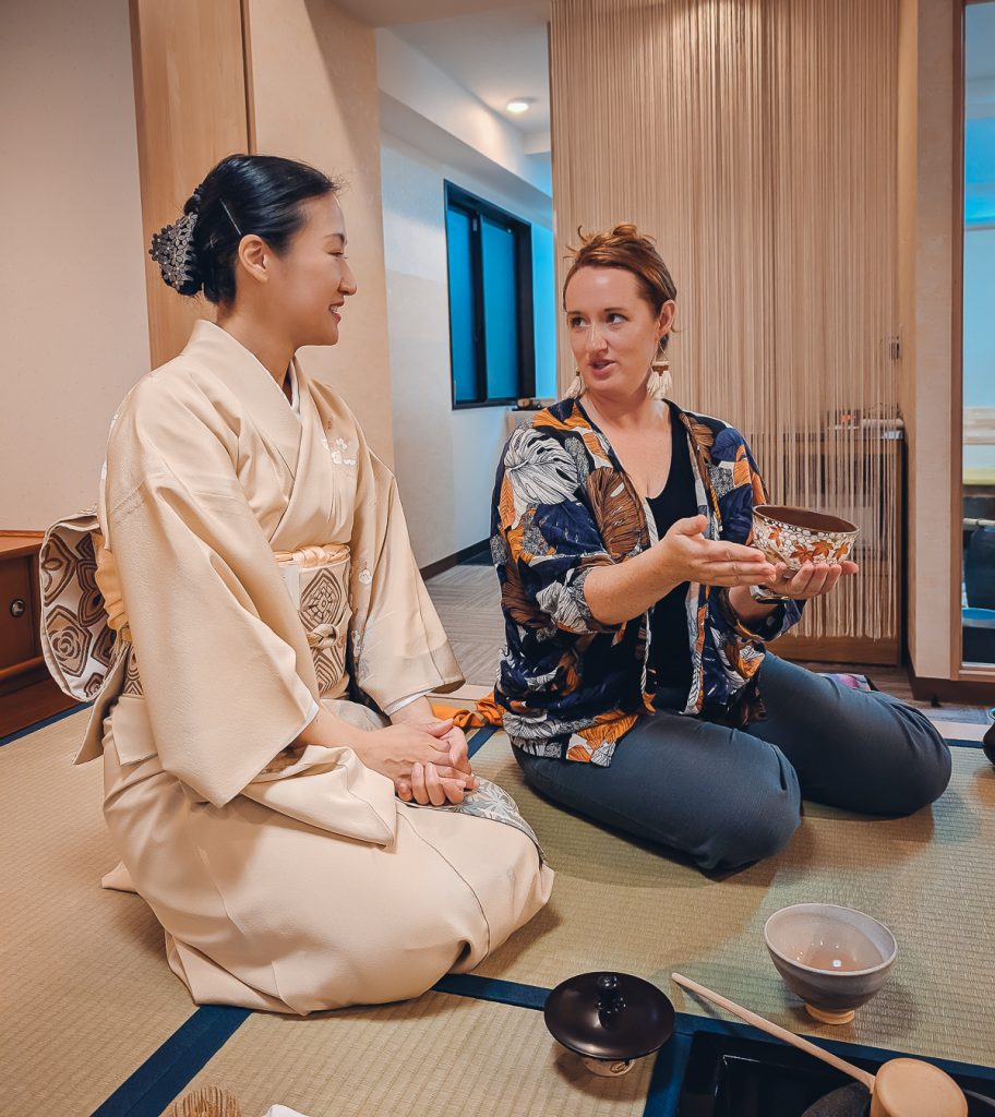 Sooz sitting on a tatami mat, holding a ceramic bowl and speaking to a Japanese woman in a kimono who is kneeling next to her.