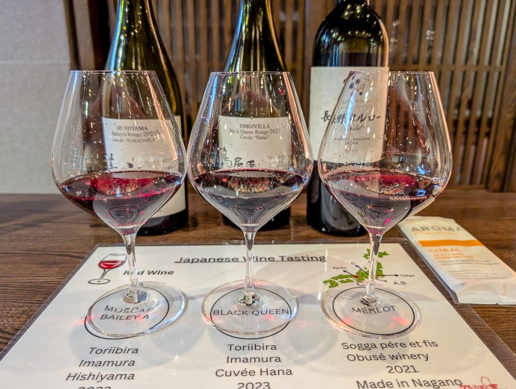 Three wine glasses with a bit of red wine in them sitting on a sheet with information about the different wines with bottles behind them and text reading "Japanese Wine Tasting."