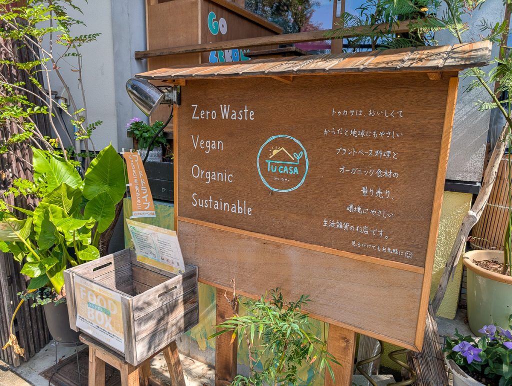 A wooden sign with "zero waste vegan organic sustainable" written in English on one side and Japanese tect on the other. In front of the sign is a wooden box with a "food box" label.