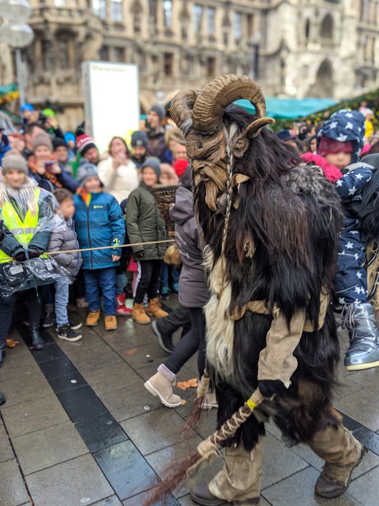 a costumed person with a mask, fur, and horns and a kid on their back depicting Krampus in Munich's Krampus Run