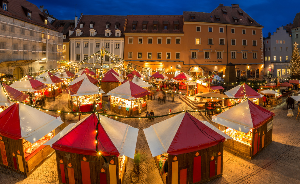 Christmas market stalls with red and white roofs in a square at night with fairy lights all over the top.