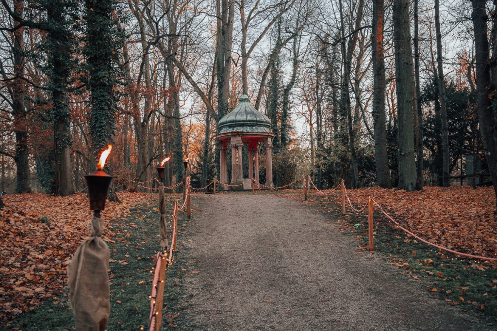 A dirt path lined with fire torches and fairy lights leading to a stone portico surrounded by trees.