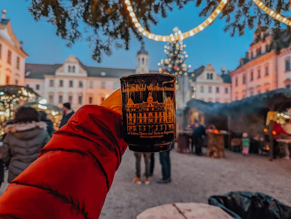 An arm in a red coat holding a Christmas mug from the Regensburg romantic Christmas market in front of an out of focus Christmas market with a lit tree.