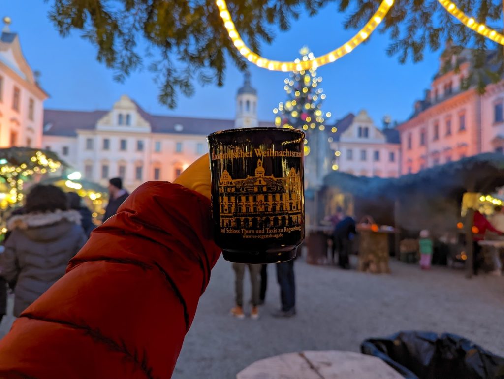 An arm in a red coat holding a Christmas mug from Regensburg in front of an out of focus Christmas market with a lit tree.