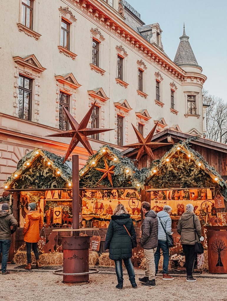 People standing in front of a Christmas market stall with a palatial building in the background.