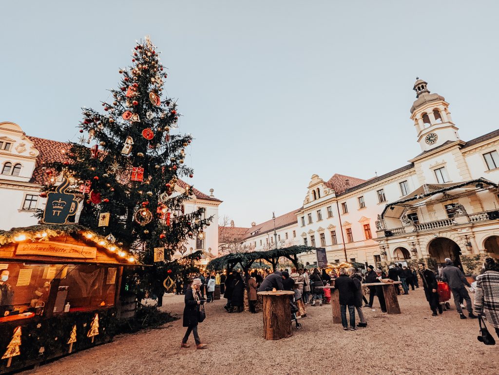 A Christmas tree, gluhwein stall and people in a square surrounded by palatial buildings.