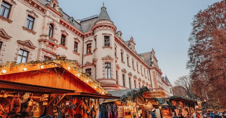 Wooden Christmas stall chalets in front of a beautiful palace with trees to one side.