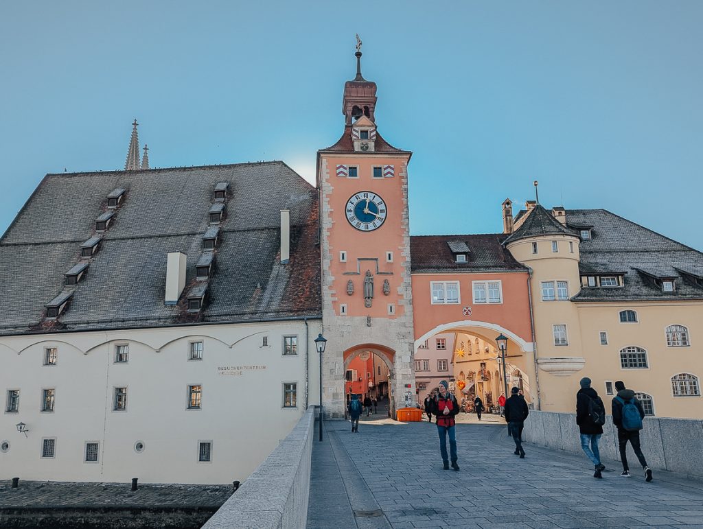 A bridge with people walking on it leading to pastel colored buildings with a clock tower and arches underneath.