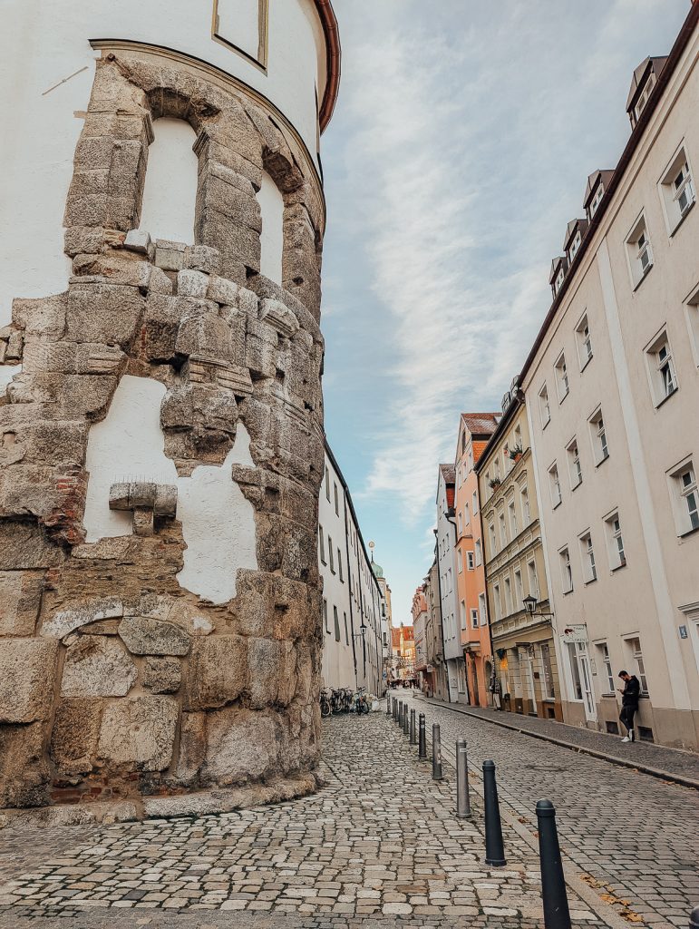 A cobbled street with light-colored buildings on one side and a rounded tower with old stones on the other.