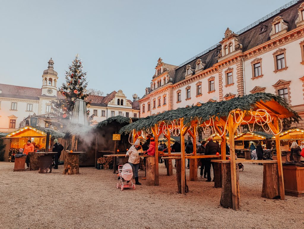 Covered tables, a Christmas tree and smoke coming from a grill in a square surrounded by a palatial building.