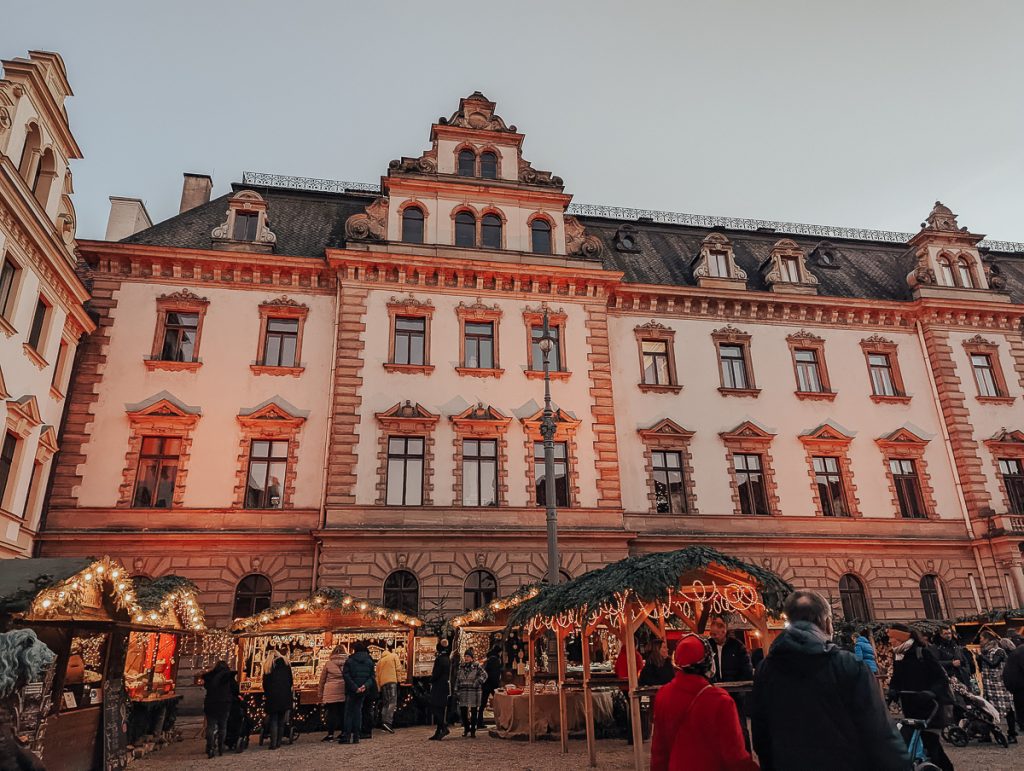 Wooden Christmas market chalet stalls in front of a palatial building.