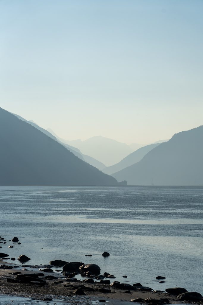 A body of water with rocks in the foreground and mountains in the background, fading into a misty sky.