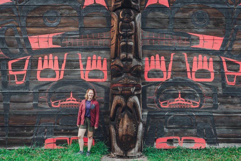 Sooz standing and smiling next to a huge wooden totem pole in front of a wooden building with red and black designs all over it.