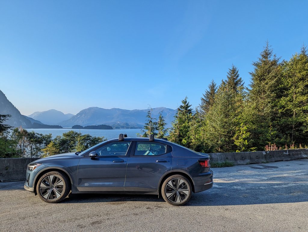A car parked on the side of a highway with water, trees and mountains in the background.