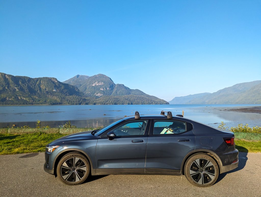 A car parked on a road next to some grass and a large body of water with forested hills and blue sky in the background.
