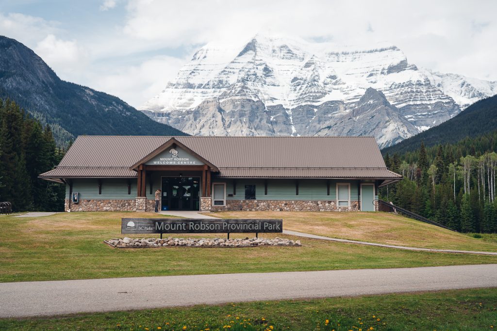 A building with "Mount Robson Welcome Centre" written above the door with a sign on the grass in front that reads "BC Parks Mount Robson Provincial Park." A rocky mountain with snow at the top rises from behind.