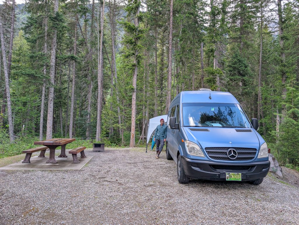 A man leaning against a campervan that parked next to a picnic table with trees in the background.
