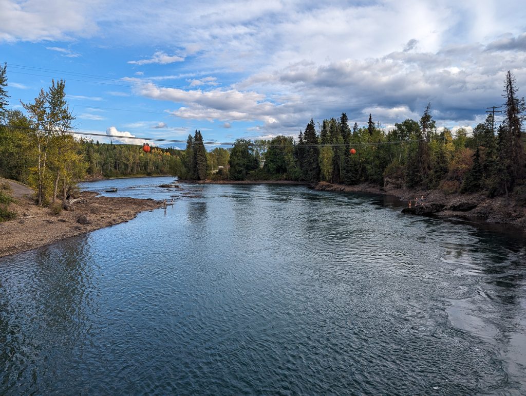 A wide river with trees on either side and power lines going above while people can be seen far away enjoying the waterside.