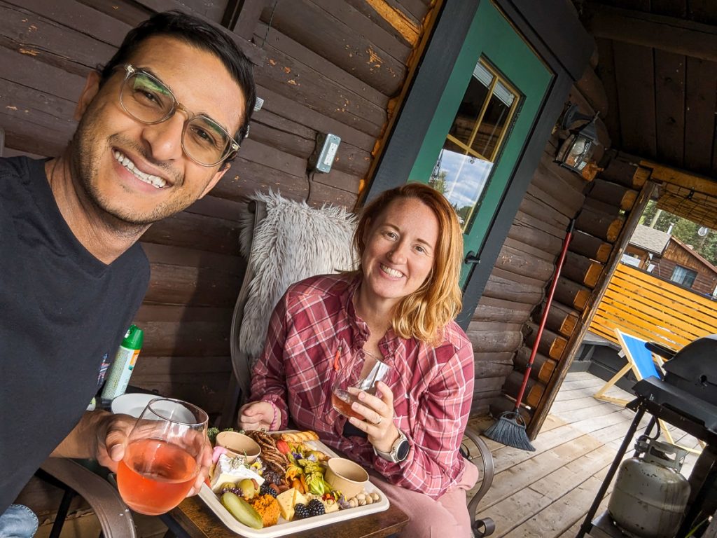Sooz and Ganesh sitting on a cabin porch with wine and a platter of food smiling at the camera.