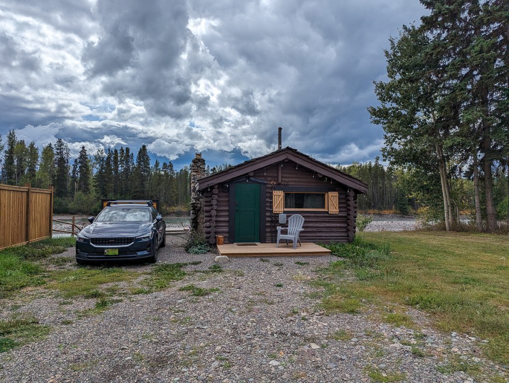 A car parked on a stone path next to a rustic wood cabin with a river and trees in the background.
